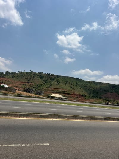 The image shows a view of the city of Kugbo, Nigeria. A highway runs along the bottom of the image. To the left of the highway, a hillside is visible, covered in trees and some buildings. Above the hillside, there is a blue sky with clouds.