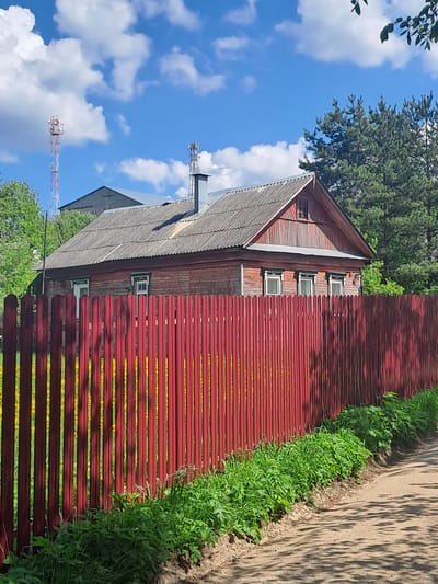 The image captures a scene in Avdot’ino, Russia, featuring a wooden house with a reddish-brown exterior and a grey, gabled roof. The house has a series of windows visible along its side. Adjacent to the house is a long, red wooden fence, constructed with horizontal planks. Green vegetation grows along the base of the fence. A dirt path runs parallel to the fence on the right side of the image, with shadows cast upon it. In the background, there is a clear blue sky with scattered white clouds. A tall telecommunication tower is partially visible in the lower left corner, positioned behind the house and amidst the trees.