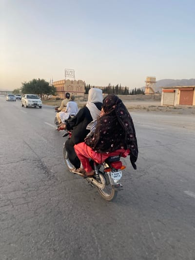 The image depicts a road scene in Spezand, Pakistan, captured from a slightly elevated perspective behind a moving motorcycle. In the immediate foreground, a silver-colored motorcycle travels away from the viewer. It carries four individuals: an adult male driver, an adult female passenger in a black abaya and white headscarf, an adult female pillion rider in a black top, red pants, and a black patterned dupatta, and a child positioned in front of the rear female passenger. The motorcycle's license plate, "MAH 9875", is visible below a red reflector, and "SPEED" is visible on the seat. The paved road surface exhibits cracks and wear.

Further down the road, to the left, another male on a motorcycle, dressed in light-colored attire, is visible. Multiple white sedans are also present on the road, moving in the same direction.

The background features a semi-arid, open environment. To the left, a line of green trees partially obscures a distinctive light-brown building with multiple arched windows and a crenellated parapet. A large sign with white Arabic script is mounted on the roof of this building, and two mosque minarets are visible behind it. To the right, the landscape is drier, featuring a tall water tower structure with a cylindrical tank. Two single-story structures with rolled-down metal shutters, possibly abandoned shops, stand near the roadside. Distant mountains form the horizon under a pale, clear sky, indicative of late afternoon or early evening light.