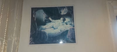 A framed painting depicting a reclining nude figure, likely Venus, rests on a white wall in what appears to be a room. The painting is oriented horizontally and is centrally placed on the wall. To the left of the painting, a sheer, cream-colored curtain hangs, partially obscuring the wall. The painting itself is rendered in a classical style, with soft lighting and a dramatic composition. The figure in the painting is light-skinned and appears to be lying on a bed or couch, draped in flowing fabric. There are other figures or elements in the background, including what looks like a cherub or angel in the upper right corner and a shadowed figure in the upper left. The wall itself appears to be textured and shows some signs of age, with small imperfections. The overall scene suggests an indoor setting, possibly a domestic space in Kandyagash, Kazakhstan.