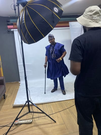 This media file captures an indoor photoshoot session in what appears to be a professional studio in Animashawun, Nigeria. The primary subject is a man, centrally positioned and posing, dressed in traditional Nigerian attire. He wears a deep blue agbada (a flowing garment) with intricate gold and black geometric embroidery on the chest, matching blue trousers, and a patterned cap (fila). He also has glasses, a gold wristwatch, a black bracelet, and holds a black walking stick in his left hand, presenting a confident demeanor.
In the foreground, partially obscuring the main subject's head, is the back of another individual, likely the photographer or an assistant. This person wears a light beige bucket hat and a black short-sleeved t-shirt.
Studio equipment is prominent. A large black softbox with golden ribs, bearing the "EDC" logo, is positioned to the left, acting as a key light source. It is mounted on a black light stand, visible with "K&F Concept" branding. A seamless white paper backdrop provides a clean background for the subject, extending onto the floor. Behind the white backdrop, several rolls of colored paper backdrops in hues like pink, blue, yellow, and red are visible against the wall. The floor on the right features light-colored wooden planks, where a black power strip with multiple outlets is placed.
The image itself is rotated 90 degrees clockwise, presenting the scene sideways. The lighting is artificial, indicative of a controlled studio environment. The time of day is not discernible.