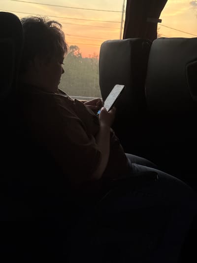 The image depicts a person in silhouette, reclining within the interior of what appears to be a vehicle, such as a bus or train, in Varvarino, Russia. The individual is seen in profile, their head resting against a seat, with light-colored, somewhat messy hair visible.

The central activity involves the person actively using a smartphone, holding it with both hands. The phone's screen is illuminated, displaying numerous lines of text, although the specific words are not discernible. The person's attention is entirely focused on the device.

The scene is strongly backlit by a large window to the left, through which a vivid outdoor view is visible. The sky outside is awash in warm, glowing orange and yellow tones, strongly suggesting either sunrise or sunset. This intense backlighting casts the vehicle's interior and the person into deep shadow, creating a striking silhouette effect. Beyond the window, indistinct forms resembling trees or distant structures can be observed against the colorful sky. The visible parts of the vehicle's interior, including sections of seats and panels, are dark and lack detail due to the shadows. The atmosphere conveys a sense of quiet travel during the twilight hours.