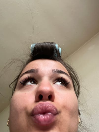 A close-up, low-angle self-portrait of a woman with dark hair and brown eyes. She is pouting her lips toward the camera. She has fair skin, dark eyebrows, and full eyelashes. A blue hair roller is visible in her hair. The background is a plain beige wall. The image was not taken in Bijedanj, Montenegro.
