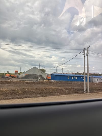 The image, taken from inside a vehicle and showing a reflection of its interior (dashboard, steering wheel, seats) on a window in the upper left, depicts an industrial or construction site under an overcast sky.

The scene is dominated by infrastructural elements and building materials. Several concrete utility poles with numerous power and communication lines stretch across the midground from left to right. To the right, a long, low blue building with multiple white windows and what appear to be air conditioning units is visible, possibly serving as an office or temporary structure.

Further back, large conical piles of grey and black crushed stone or gravel are prominent. Several orange dump trucks are positioned around these material piles, some appearing to be in use or waiting, with one seemingly loaded. In the distance, an electrical transmission tower stands against the cloudy horizon, and other smaller buildings or structures are faintly visible. The ground throughout the visible area is largely undeveloped, consisting of disturbed earth and dirt.

The sky is heavily clouded, suggesting an overcast day with no direct sunlight. No people are clearly visible, but the presence of machinery and prepared materials indicates ongoing or recent industrial activity. There is no discernible text in the image. The setting appears to be an undeveloped area undergoing construction or serving as a material depot, consistent with an industrial or infrastructure project in Kabanikha, Russia.