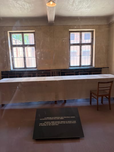 This image depicts an interior scene, likely within a historical site or museum, given the presence of informational text. The room appears sparsely furnished with two windows, each divided into four panes. The walls are a neutral, textured color, and a simple wooden chair and a long, covered table are visible. The lighting suggests it could be daytime, with natural light entering through the windows.
A prominent feature is a dark plaque with text inscribed in both Polish and English. The text describes the room as a place where summary sessions of a Gestapo court were held, and the most frequent sentence was the death penalty by shooting at the "Death Wall." This suggests the location is a site of historical significance related to World War II and the Holocaust, likely Auschwitz-Birkenau, given the context provided.
The overall atmosphere is somber and reflective, conveyed through the starkness of the environment and the historical information presented. The image is oriented vertically, with the top of the scene on the left. No people are visible in the frame.