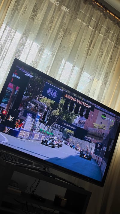 The image, captured in the city of Rogachevo, Bulgaria, displays an indoor setting with a television screen as the focal point, positioned against a brightly lit window. The perspective is slightly angled and tilted, looking down at the television and the elements behind it.

A black Samsung flat-screen television occupies the lower and central part of the frame. The screen is active, broadcasting a motor race event, identified by text in the upper left corner as the "GRAND PRIX OF MONACO 2020." The race appears to be a Formula E event, with multiple open-wheel electric race cars visible on an urban street circuit. Trackside elements include prominent "LOUIS VUITTON" branding on overhead gantries and barriers, the "FIA" logo on an overhead structure, and additional branding such as "TAG HEUER" partially visible. Race officials in high-visibility safety gear and spectators are observable in the background of the televised race scene. A digital display on the television's lower frame shows "16:03." The broadcast channel "DIEMA SPORT 3 LIVE" is visible in the upper right corner of the screen.

Beneath the television, a dark media console or shelf holds several objects. These include a bundle of black cables, a dark remote control, a small black electronic device, and a dark cylindrical container or candle. A stack of books is visible on the left side of the shelf.

In the background, behind the television, light-colored, semi-sheer curtains with a distinct floral or patterned lace design are visible. These curtains are illuminated by strong natural light emanating from a window, suggesting a daytime scene. The curtains are suspended from a gold-colored or wooden curtain rod, secured by rings, visible in the upper right corner of the background.