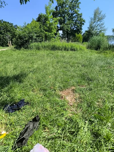 The image depicts a grassy area in Chézenas, France, under a clear blue sky with scattered trees in the background. The ground is covered in green grass, with a patch of bare earth visible in the middle ground. A black garment, possibly clothing, is lying on the grass in the foreground, along with some other indistinguishable objects. In the background, there's a line of trees and vegetation, with glimpses of what appears to be a body of water on the right side. The scene gives the impression of a park or open space. No people are visible in the frame.