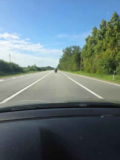 The image is a first-person perspective from inside a vehicle looking out at a two-lane asphalt road. The sky is clear blue with scattered white clouds. On the right side of the road, there is a dense line of green trees. On the left side of the road, there are also trees and some vegetation, with a telephone pole visible in the distance. The road itself has clear white lane markings. In the distance, two motorcycles are traveling in the same direction as the viewer's vehicle. The foreground of the image shows the dashboard of the vehicle. The overall scene depicts a daytime drive on a rural highway.