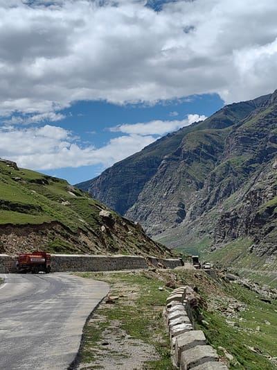 In Yari-Khoksar, India, a road winds through a mountainous landscape under a partly cloudy sky. The road, paved with asphalt and bordered by a low stone wall on the right, curves to the left in the foreground. To the left of the road, a grassy, rocky slope rises steeply, while on the right, another, steeper mountain face is visible, its upper reaches covered in greenery and its lower sections rocky and barren.

A red tanker truck, its rear facing the camera, is parked on the side of the road on the left. Further up the road, a dark-colored truck and a white vehicle are visible, suggesting ongoing traffic or construction activity. The mountains themselves are rugged and imposing, with shadows indicating the direction of the sun. The overall scene depicts a remote mountain pass, likely a transportation route through the Himalayas.