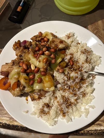 A close-up, overhead shot displays a meal served on a white circular plate, likely taken indoors. The plate occupies the central and right portions of the frame, resting on a table with a dark, patterned surface on the left and a lighter, possibly wooden, textured surface on the right.

The meal consists of a generous serving of white rice, partially covered with a dark, savory sauce or seasoning, and a hearty mix of other ingredients. A metal fork is visible, inserted into the rice, suggesting the meal is either about to be eaten or is in progress. The main dish, located on the left side of the plate, features chunks of cooked meat (possibly chicken), slices of orange carrots, red onion pieces, and what appears to be a soft, light green vegetable (perhaps zucchini or bell pepper), all covered with melted cheese. Numerous roasted peanuts are sprinkled generously over the entire main dish, adding a textural element.

In the background, on the far left, a light green circular container or lid is partially visible. In the lower-left corner, a black Duracell Ultra battery is lying on its side. The lighting appears to be artificial, casting some shadows. The scene depicts a prepared meal, ready for consumption. There are no specific visual cues within the image to directly confirm the location as Marra, India, but the presence of peanuts is common in various cuisines, including Indian. The visible text on the battery is "DURACELL ULTRA".
