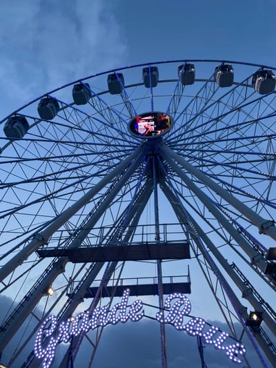 An upward-angled photograph, tilted slightly to the left, captures a large, illuminated Ferris wheel against a blue-grey, cloudy sky. The lighting suggests it is either dusk or early evening. The Ferris wheel's intricate metal structure features numerous spokes radiating from a central hub, all adorned with countless small blue and white LED lights that outline its form. Several enclosed passenger gondolas are visible along the left side of the wheel's circumference. At the very center of the wheel, a circular LED screen displays a dynamic, colorful video or image, though its specific content is not clearly discernible. To the right of the Ferris wheel, a prominent illuminated sign, also made of individual white bulbs, spells out the word "Roulette" in a decorative, cursive script. Supporting metal framework for the ride is also visible on the right side of the image. The scene depicts a vibrant amusement park or fairground attraction, located in Luik, Belgium.