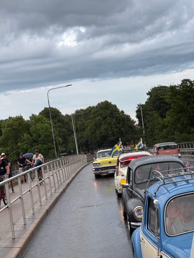 A convoy of vintage cars, several adorned with Swedish flags, drives down a wet bridge in Falkenberg, Sweden. The sky is overcast, suggesting rain. People on bicycles are visible on the sidewalk to the left of the cars. The cars include a yellow classic car, a red station wagon, a black Volkswagen Beetle, and a blue and cream Volkswagen Beetle in the foreground. The bridge has a metal railing along the pedestrian walkway. Trees line the background.