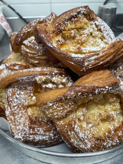 A close-up shot showcases four golden-brown, square-shaped pastries, likely danishes, arranged on a metal tray. Each pastry features a flaky, layered exterior dusted with powdered sugar. The center of each pastry is filled with a crumblike topping, and a hint of a yellow-colored filling is visible beneath the topping. The pastries are positioned close together, creating a visually appealing display.
