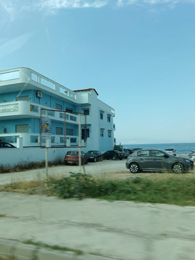 The image depicts a blue building located in the Municipal Unit of Sikyona, Greece, situated near the sea. The building has multiple levels with balconies and windows. In front of the building, a parking area is visible with several cars parked, including a gray hatchback, a red hatchback, and a black sedan. The sea is present in the background, with a hazy landscape visible on the horizon. The sky is clear with some wispy clouds. The foreground shows a paved area and some vegetation, suggesting a roadside or public space. The image appears to be taken from a moving vehicle, as indicated by motion blur in the foreground.