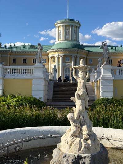 This is a landscape-oriented, slightly tilted, daytime outdoor photograph featuring a grand, yellow building with a green roof and white architectural details. The building appears to be a palace or a large estate, with multiple windows and classical statues adorning its exterior. In the foreground, a stone fountain with sculpted figures is visible, with water gently splashing. Lush green vegetation grows around the fountain and the building. The sky is a vibrant blue with scattered white clouds.
The scene depicts a tranquil moment, likely in the late morning or early afternoon, given the bright sunlight casting shadows. The weather appears to be clear and pleasant. The overall impression is one of elegance and historical grandeur. The location is identified as Arkhangel'skoe, Russia, which is known for its beautiful estate and palace.
While there are no discernible text elements in the image, the presence of people, though small in the frame, suggests the location is accessible to visitors and is likely a tourist attraction or a well-maintained public space. The statues in the fountain depict figures in dynamic poses, adding a sense of artistry and movement to the scene. The architectural style of the building is reminiscent of European classical or baroque designs.