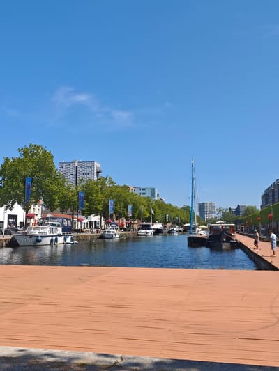 The image depicts a sunny day in the Spoorzone district of Tilburg, Netherlands, specifically along the canal that runs through the area. In the foreground, a large, flat, orange-colored platform made of narrow planks is visible, extending across the lower portion of the frame. This platform appears to be a temporary structure.

Beyond the platform, the canal is filled with a variety of boats. On the left side, several houseboats and yachts are moored along a stone embankment. These boats vary in size and design, with some featuring white hulls and blue or grey coverings. Further down the canal, a tall, slender sailboat with a blue and white sail is anchored. On the right side of the canal, a long, brown wooden pier juts out into the water. Two individuals are standing on this pier, appearing to fish.

The background reveals a mixed urban landscape. Lush green trees line the canal on both sides, interspersed with buildings of various architectural styles. On the left, a white building with a "FITNESS" sign is visible, along with taller, modern apartment buildings. Flags, some blue with white designs, are displayed along the waterfront. On the right, more modern buildings with dark facades and balconies overlook the canal. The sky is a clear, bright blue, indicating good weather.