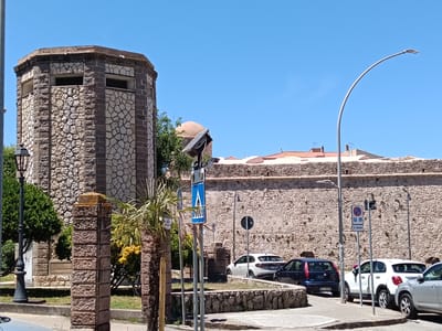 The image depicts a street scene in Alghero, Italy, likely near a historic fortification. In the foreground, a stone pillar and a palm tree are visible. To the left, an octagonal stone structure with windows rises against a clear blue sky. Behind it, a large stone wall, characteristic of historic city defenses, stretches across the background. Several cars are parked along the street, including a white SUV, a dark blue hatchback, and a silver sedan. Street signs, including a pedestrian crossing sign and a parking sign with a wheelchair accessibility symbol, are present. The overall environment suggests a sunny day in a Mediterranean city.