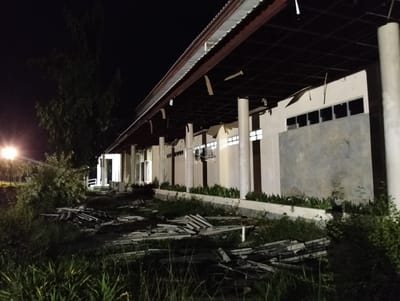 This media file captures an abandoned or under-construction building at night in Palohgadeng, Indonesia. The scene is dimly lit by artificial light sources, including a street lamp visible on the left, casting long shadows and creating a somewhat eerie atmosphere. The building itself appears to be a long, open-sided structure with a corrugated metal roof supported by numerous large, cream-colored concrete pillars. The underside of the roof structure is visible, showing a dark grid of beams and some scattered debris.
The walls of the building are constructed from a combination of light-colored concrete panels and what appear to be large windows, many of which are dark or boarded up, suggesting disuse. A row of horizontal windows runs along the upper part of the wall. Hanging from the roof structure are pieces of wood and other materials, possibly remnants of construction or damage.
In the foreground, the ground is covered in overgrown grass and weeds. Piles of discarded construction materials, including what looks like broken roof tiles and planks of wood, are scattered across the area, further emphasizing the abandoned or unfinished state of the site. A large tree with dense foliage is visible on the left, silhouetted against the dark night sky. The overall impression is one of neglect and decay, set against a quiet, dark environment. No people or animals are visible in the image. There is no visible text.