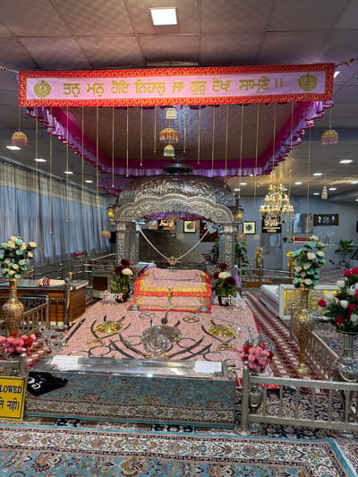 The image depicts the interior of a Gurdwara, a Sikh place of worship, likely in Chumkar, India, given the Punjabi script visible on the banner. The central focus is a raised platform, possibly an altar, covered with pink and gold fabric. On this platform rests a large, ornate silver canopy. Several decorative elements adorn the space, including an elaborate chandelier hanging from the ceiling, floral arrangements in vases, and patterned carpets covering the floor. A prominent banner with Sikh religious text in Punjabi and a Khanda symbol is displayed above the canopy. The scene is richly decorated with ornate metallic objects and intricate textiles, suggesting a place of significant religious importance and community gathering. There are no people visible in the frame.