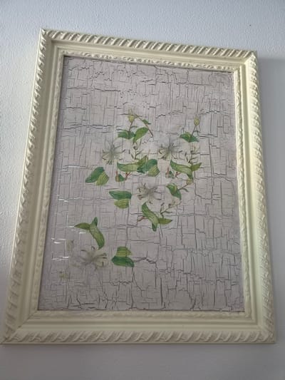 This is a photograph of a framed picture hanging on a textured, light-colored wall. The picture itself features a floral design against a distressed, light gray background that is heavily cracked, resembling aged paint or plaster. The artwork depicts several delicate white flowers with green leaves and stems. The frame is wide and appears to be made of a light-colored material, possibly painted wood, with a decorative rope-like or braided pattern along its outer edge and a simpler patterned border on the inner edge. The lighting suggests it is daytime, and the image is taken from a slightly elevated angle. There is no visible text in the image. The setting appears to be indoors, in a domestic or possibly a small office space, given the simple wall and the framed artwork. The overall impression is one of rustic or vintage decor.