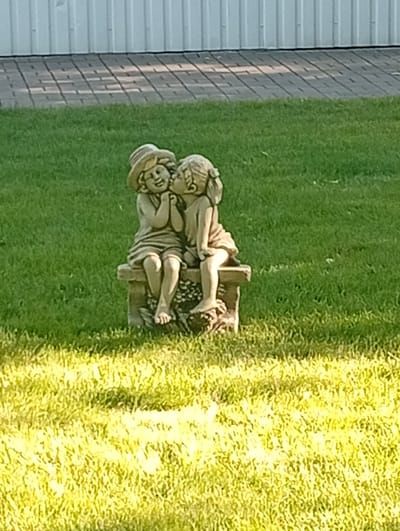 The image shows a statue of two children sitting on a bench in a grassy area. One child is wearing a hat and the other child is kissing them. The statue is made of stone and appears to be old. In the background, there is a white building with a corrugated metal siding, and a paved area with a checkered pattern. The scene is illuminated by sunlight, casting shadows on the ground.