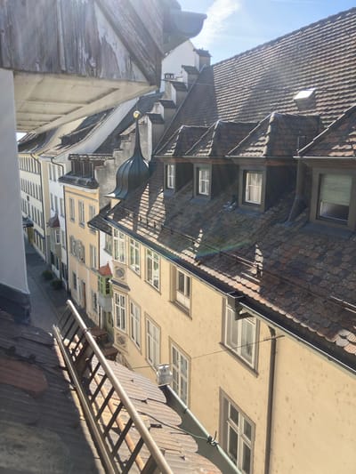 The image, originally rotated 90 degrees counter-clockwise, depicts a high-angle view looking down into a narrow street or alley lined with historic buildings in Schaffhausen, Switzerland. The scene is bathed in bright daylight, suggesting late morning or early afternoon.

Dominating the upper portion of the view are steeply pitched, dark tiled roofs, likely made of terracotta or slate, punctuated by numerous dormer windows. Below these, the building facades are predominantly light-colored, in shades of creamy yellow, beige, and white, featuring many rectangular windows with white frames. A distinctive building in the mid-ground boasts a dark, ornate spire or cupola. Another building exhibits a rustic, weathered wooden gable end.

In the foreground, a lower, dark tiled roof is visible, where a metal ladder rests horizontally. Utility lines or cables can be seen stretching between buildings. Direct sunlight, originating from the top-left (when corrected), casts sharp shadows and highlights the textures of the roofs and walls. A faint lens flare or rainbow effect is present in the sunlight. The visible sky is clear and blue with a few scattered clouds. The scene is devoid of human activity, conveying a sense of quietude in this historic European urban setting. No visible text is present.