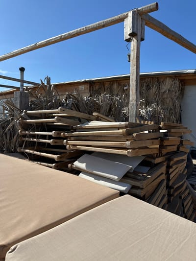 The image displays an outdoor scene, characterized by a clear, bright blue sky in the upper left portion. The perspective is upward, revealing a structure crafted from sturdy, rustic wooden posts and beams. This wooden framework supports a roof or awning made of dried palm fronds or similar natural thatch material, contributing to a tropical or coastal aesthetic.

A significant part of the image, particularly to the right and in the mid-ground, is occupied by numerous wooden lounge chairs or deck chairs. These chairs are neatly stacked, suggesting they are either in storage or being organized for use or transport. Several chairs are equipped with light-colored, possibly beige or white, fabric cushions.

Notable details include a small black object, resembling a security camera or sensor, mounted on one of the prominent wooden support posts on the left. Below this, a black-capped pipe or vent extends upwards.

The scene is illuminated by strong daylight, indicating clear and sunny weather. There are no individuals present, and no active event is captured, giving the impression of a tranquil or utilitarian setting. The overall style of the wooden construction and furniture points to a casual outdoor environment, typical of a beachside establishment, resort, or open-air café. This is consistent with the provided location context of Anatoliki Attiki, Greece. No visible text is present in the image.