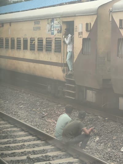 The image depicts a dusty railway scene, likely in Seoni Malwa, India, with a pale yellow train stopped on tracks. The train has "SLEEPER" written on its side in blue English lettering and Hindi script above it. A man in a white shirt and light blue pants stands in the open doorway of a train carriage, his hand near his mouth as if speaking. Below the train, on the gravelly tracks, two men are seated. One is wearing a light green t-shirt and the other a dark olive green t-shirt and dark pants, his arm bent as if holding something. The atmosphere is hazy, suggesting dust or smoke from the train or its surroundings. The railway tracks are composed of ballast and sleepers, with visible fastening mechanisms. The train carriage features multiple barred windows.