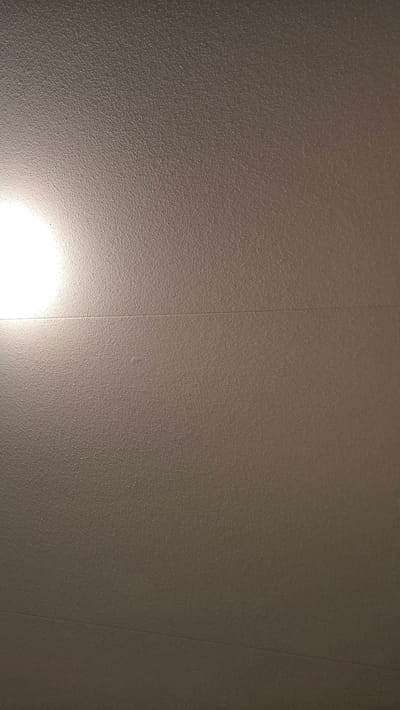 The image shows a textured, light brown ceiling. A bright light source is visible on the left side of the frame, casting a warm glow across the surface. A faint horizontal line, possibly a seam or joint, is visible across the middle of the ceiling. The texture appears to be consistent across the entire visible area, suggesting a uniform material like painted drywall or plaster. There are no people, objects, or specific environmental features visible beyond the ceiling itself and the light source. The image is a close-up shot of the ceiling, with no context provided to determine its location within Vlaardingen, Netherlands.