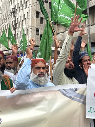 A group of men are gathered outdoors, holding green flags and a large banner. The flags are attached to wooden poles and some display white lettering. The men are wearing traditional Pakistani clothing, including shalwar kameez and caps. One man has a prominent grey beard and wears a light blue shirt and an orange cap. Another man, wearing a dark blue shirt and a blue cap, is also visible, along with several other individuals. The atmosphere appears to be that of a rally or protest, with many hands raised in the air. Some men are smiling and appear engaged in the event. In the background, a multi-story building with many windows is visible. A large, cream-colored banner is partially obstructing the view, with some text visible on a white strip at the top. The visible text on the banner and flags appears to be in Urdu. The scene is outdoors, likely during the daytime. The overall mood suggests a political or religious gathering.