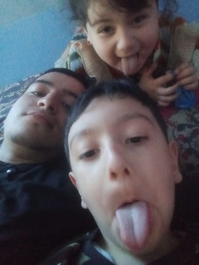 A close-up, indoor photograph, likely a selfie, depicts three individuals in Kasan, Uzbekistan. In the lower left quadrant, an adult male, appearing to be in his late teens to early thirties with short dark hair and faint facial hair, is positioned with his head tilted slightly upwards, looking towards the camera with a neutral expression. He wears a dark-colored shirt. In the foreground, occupying the lower right and central portions, is a male child, estimated to be between 6 and 10 years old, with short dark hair, looking directly at the camera with his tongue extended. He is wearing a dark, possibly patterned, collared shirt. Above and partially behind the older child, and to the right of the adult male, a younger female child, approximately 2 to 4 years old, with dark hair, is visible. She is also sticking her tongue out, smiling, and wearing a patterned garment. A light blue surface is visible in the upper left background, suggesting a wall or fabric, while other blurred patterned fabrics are present elsewhere, indicating an indoor setting.