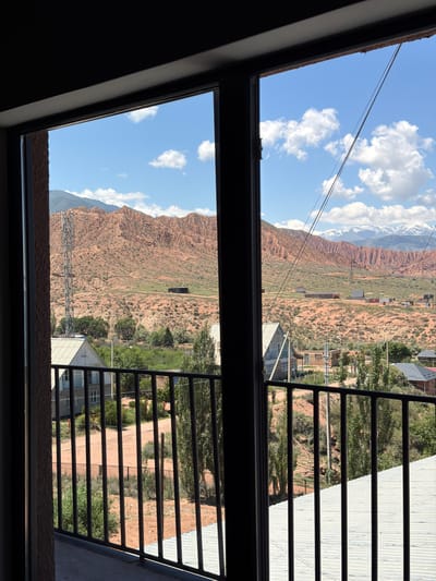An outdoor view from a balcony in Kaji-Say, Kyrgyzstan, looks out onto a valley with red, rocky mountains in the background. The sky is blue with scattered white clouds. In the middle ground, there are several small buildings, some with metal roofs, and trees. A tall communication tower stands on the left side of the image. In the foreground, a black metal railing is visible. The overall scene depicts a rural or semi-rural area with mountainous terrain.