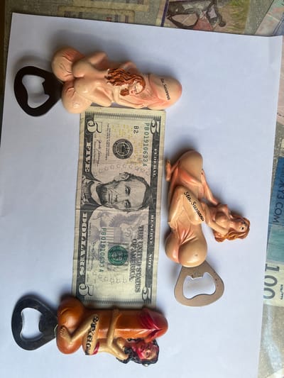 The image displays three novelty bottle openers shaped like nude figures. One bottle opener is positioned horizontally across a five-dollar bill, featuring a figure with red hair and the inscription "Sta. Susanna." Another similar bottle opener is located to the right of the five-dollar bill, also bearing the "Sta. Susanna" inscription. A third bottle opener, oriented vertically, is at the bottom of the frame, featuring a figure with darker hair and red clothing, with the word "Barcelona" visible on its torso. The background consists of a white surface with various foreign banknotes scattered around it. There is no indication of the city of Schwäbisch Hall, Germany, in the image.