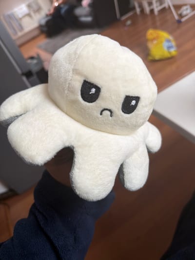 A cream-colored reversible octopus plush toy, displaying an upset expression with embroidered black eyes and a downturned mouth, is held in the foreground. The underside of its eight tentacles reveals a grey fabric. A human hand, partially visible and clad in a dark blue long-sleeved garment, holds the plush from below. The background depicts a blurred indoor domestic environment. A light brown wooden floor is prominent, with a crumpled yellow plastic bag situated on it to the right. A dark sofa or seating arrangement is visible in the left background, with the legs of an individual in dark trousers resting on it. A light-colored rug is beneath the sofa, and a white or light-colored table surface is present on the far right.