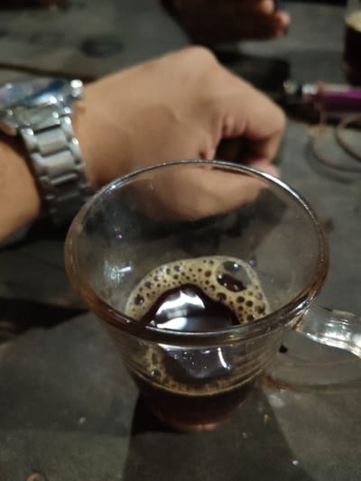 The image displays a close-up view of a clear glass mug containing a dark brown beverage, likely coffee or tea, topped with a layer of foam and bubbles, some exhibiting iridescent qualities. This scene is situated in Balla, Bangladesh. The mug rests on a dark, textured surface. To the upper left, a human forearm with light skin is visible, adorned with a silver-toned metal link watch on the wrist. In the blurred background, another indistinct hand or arm is partially present, alongside a blurred object with metallic and purple elements. The environment is dimly lit, consistent with an indoor setting under artificial illumination. No direct actions are depicted, but the arrangement suggests a paused moment of beverage consumption.