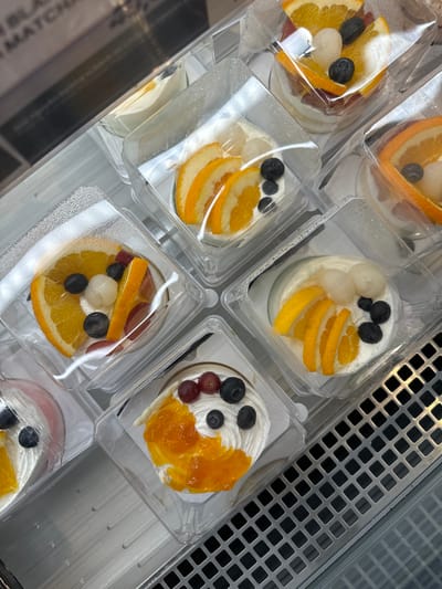 In Phu Thuy, Vietnam, a retail display case showcases several individually packaged desserts. The desserts, presented in clear plastic containers, are topped with white cream and adorned with various fruits such as orange slices, blueberries, grapes, lychees, and what appears to be apricot jam. The display case itself has a metallic grid at the bottom, and reflections are visible on the glass enclosure, obscuring some of the background. The lighting suggests an indoor setting, likely a bakery or dessert shop. No people are visible in the image.