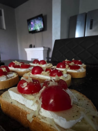 The image shows a close-up of several bruschetta appetizers arranged on a dark surface. Each appetizer is made with a slice of baguette topped with cream cheese, cherry tomatoes, and grated cheese. The bruschetta are seasoned with black pepper. In the background, a wall-mounted television displays a colorful, abstract image. To the right of the television, a white rectangular object, possibly a shelf or part of a fixture, is visible. The overall setting appears to be indoors, with a focus on the food preparation or presentation. The lighting suggests an artificial light source.