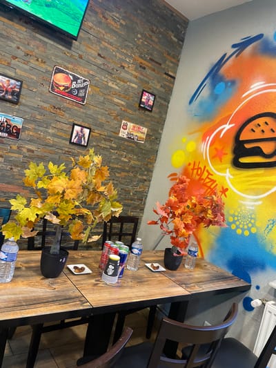The image depicts an interior scene within a casual eatery in Holtzheim, France, specifically a dining area devoid of people. The environment features two distinct wall treatments: the left wall is clad with horizontal, multi-toned stone veneers, while the adjacent right wall is painted a light grey with a vibrant, multi-colored graffiti mural.

Mounted on the stone wall are various decorative items: a flat-screen television displaying a green landscape is positioned high on the wall. Below it, a large "HOT BURGERS FAST FOOD" metal sign with a burger graphic is visible. Surrounding these are smaller framed pictures, the content of which is largely indistinct, along with a "ARIZONA US Route 66" metal sign featuring a classic car, and a license plate-style sign.

The graffiti mural on the right wall showcases a prominent black outline of a hamburger with sesame seeds, overlaid on a dynamic background of yellow, orange, and blue spray paint. White outlines, stars, and the word "HOLTZA" in graffiti script are also visible.

In the foreground, a rustic wooden dining table, comprising at least two joined sections, occupies the central space. Several dark-stained wooden chairs with slatted backs are positioned around the table.

Arranged on the tabletop are various objects:
- Two clear plastic bottles of still water are placed at either end of the visible table surface.
- Two black potted artificial plants with autumn-colored leaves act as centerpieces; one features yellow and green foliage, and the other displays red and orange leaves.
- Two small white square dishes, each containing two irregular-shaped brown food items (possibly dates or small pastries), are situated near the potted plants.
- A cluster of beverage containers is visible, including several red energy drink cans, a silver beverage can, and a stack of black and white disposable "TO GO" coffee cups.

No individuals are present in the scene, and no specific actions or interactions are depicted. The setting appears ready for customers or recently vacated.
