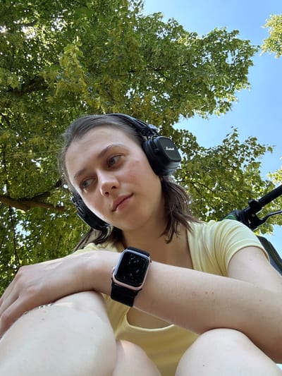 A woman is wearing headphones and a smartwatch, sitting outdoors under a tree. The sky is blue and the sun is shining. She is looking to the side and appears to be relaxed. The image was taken in Erlenbach am Main, Germany.