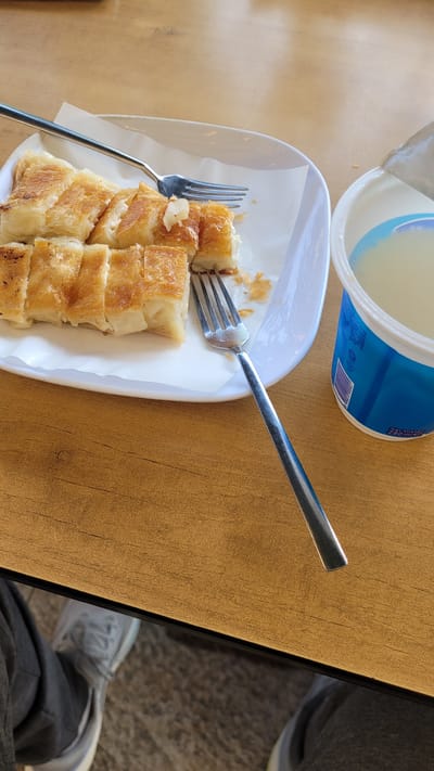 A high-angle shot shows a meal set on a wooden table. The meal consists of a sliced pastry dish served on a white plate lined with white parchment paper, two forks, and a container of a liquid substance.
The pastry, golden brown with a layered appearance, is neatly sliced into small portions on the plate. Two silver forks are placed on the plate, one directly piercing the pastry and the other resting along the side.
A cylindrical container with a white interior and blue exterior sits near the plate. The contents appear to be a creamy, light-colored liquid.
The scene is set on a wooden table with a distinct grain pattern. To the right, a portion of a person's lower body is visible, clad in gray pants and white sneakers.
The image was taken in Beylikdüzü, Turkey.