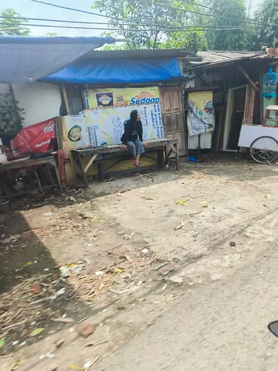 The image shows a street scene in Sukasari, Indonesia, located at latitude -6.837 and longitude 107.598. A young woman wearing a black shirt and dark pants sits on a wooden bench in front of a small shop. The shop has a blue awning and is adorned with advertisements for various products, including a brand of instant noodles called "Sedap." There are other shops and buildings in the background, and a few pieces of trash are visible on the street. The woman is looking down at her phone, and the scene appears to be an ordinary daily moment in Sukasari.