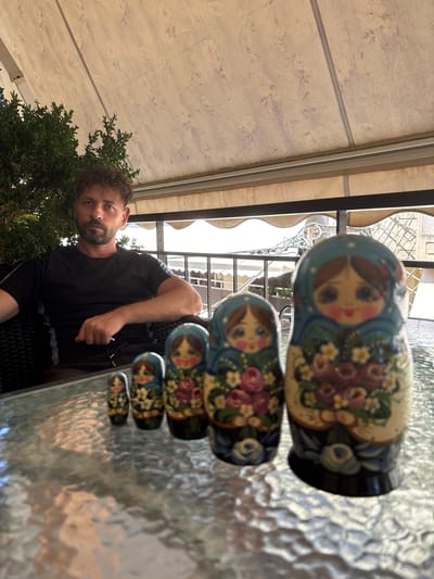 An adult male with curly, dark hair and a beard is seated in a dark chair, wearing a black t-shirt. He is looking directly at the camera. In the foreground, on a reflective glass table, are several Matryoshka dolls, arranged in a line from smallest to largest. The dolls are painted with floral patterns and have traditional facial features. Behind the man, there is a lush green plant and an awning with a floral pattern. The background suggests an outdoor seating area, possibly a cafe or restaurant, with railings and buildings visible. The overall scene appears to be outdoors in Ilion, Greece.