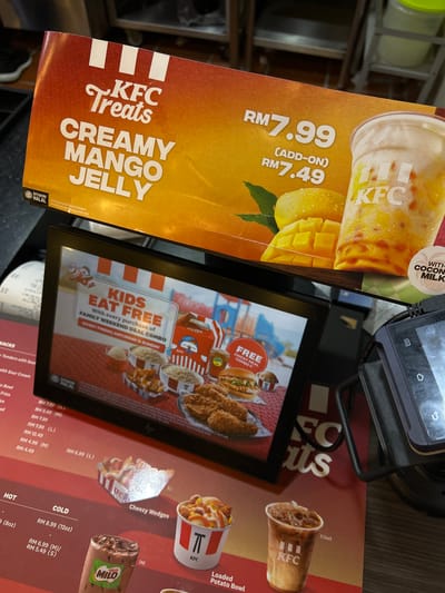 The image shows KFC promotional materials in Putrajaya, Malaysia. On the left is a poster advertising "KFC Treats Creamy Mango Jelly" for RM7.99 or RM7.49 as an add-on. The poster features an image of the drink, with the text "WITH COCONUT MILK." A digital display shows an ad for "KIDS EAT FREE" with the purchase of a "FAMILY WEEKEND DEAL COMBO" on Fridays, Saturdays, and Sundays. Next to the display is a KFC menu section with items like Loaded Potato Bowl, Cheezy Wedges (RM 6.99 L), and Milo (RM 6.99 M / RM 5.49 S). A black handheld device sits near the top of the image. The overall scene is of promotional materials and menus, likely within a KFC restaurant setting. The "DIYAKINI HALAL" logo is visible on multiple items.