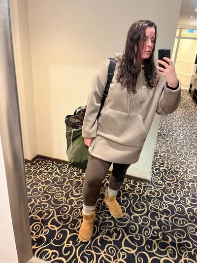 A person takes a mirror selfie. They have long, dark, curly hair and wear a beige fleece hoodie with a drawstring, brown leggings, and tan UGG boots with grey socks peeking out. A black strap crosses their body, connected to a green bag. The carpet is patterned with black swirls on a tan background. The wall behind the mirror is plain white.