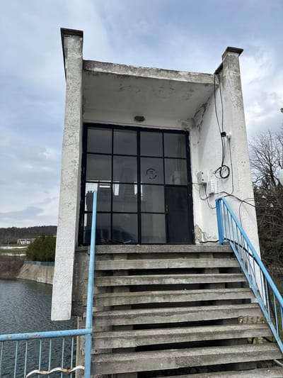 This image, captured in Баня, Bulgaria, depicts a two-level, light-colored concrete structure positioned at the edge of a body of water under an overcast sky. The building exhibits signs of weathering, including peeling paint and discoloration on its exterior. Its facade features a large, black-framed glass section comprising multiple panes, with a circular emblem visible on one pane, adjacent to a smaller solid black door on the right. An overhead light fixture is installed on the underside of the concrete overhang directly above the entrance. The right exterior wall displays several white electrical boxes and exposed black wiring running vertically and horizontally.
Access to the entrance is provided by a set of approximately 10-12 concrete stairs, each side equipped with blue metal handrails. The left handrail descends towards the water, bordering the edge of the visible structure. The dark, calm body of water, likely a reservoir, extends to a concrete retaining wall on its opposite bank. The background reveals a distant treeline, hills, and several indistinct buildings beyond the water on the left. Bare deciduous trees are visible on the right side of the frame, suggesting a late winter or early spring season.