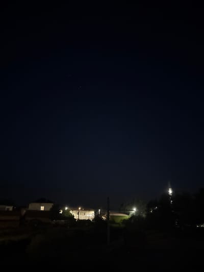 The image depicts a nocturnal scene in the city of Blatska, Bulgaria. The sky is a deep, dark blue, indicating nighttime, and a few faint stars are visible. In the lower portion of the frame, several illuminated buildings are scattered amongst dark trees and vegetation. One building on the left has a single brightly lit window. To its right, another building is partially illuminated by streetlights or internal lighting. Further to the right, more structures are visible, some with lights on. The overall impression is of a quiet, residential area at night, with the lights of homes and possibly streetlights providing the only illumination against the dark sky and landscape.