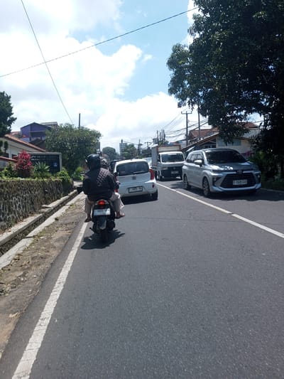 A street scene showing multiple vehicles. The focal point is a person riding a motorcycle, with a passenger on the back. To the right are several other cars, including a white Kia, a silver Toyota, and a white truck. The motorcycle license plate reads "F 5690 HBN 08/30". The Kia's license plate reads "F 1825 DL", and the silver Toyota's license plate reads "B 1528 CIU". The left side of the frame shows a raised sidewalk with a stone wall and greenery. There are power lines in the sky, a partially cloudy sky, and trees in the background. The location is not in Kioumaknao 2, Laos. Instead, the license plate indicates this is likely in Indonesia.