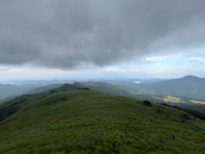 The photograph captures a landscape in Illukkumbura, Sri Lanka. Rolling green hills dominate the foreground, covered in dense grass. In the distance, a series of layered mountains fade into the hazy atmosphere. A body of water can be seen between the mountains. Above, a cloudy, overcast sky fills the upper portion of the frame, suggesting a cloudy day.