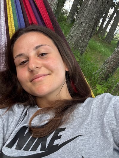 The image shows a young woman lying in a hammock outdoors, with trees and green grass in the background. She is wearing a gray sweatshirt with a black Nike logo. Her hair is long and brown, and she is looking at the camera with a slight smile. The hammock has colorful stripes. The location is specified as Kosovo, Bulgaria.