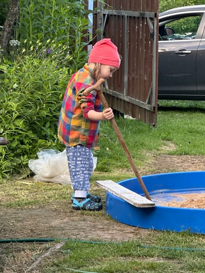 A young child wearing a red beanie, a colorful plaid shirt, and blue patterned leggings is standing in a backyard near Piriutino, Russia. The child is holding a wooden stick, which is resting on the edge of a blue sandbox filled with sand. A piece of wood is also placed on the edge of the sandbox. In the background, there is a wooden gate, a dark-colored car, and lush green vegetation. The ground is covered with grass and dirt.